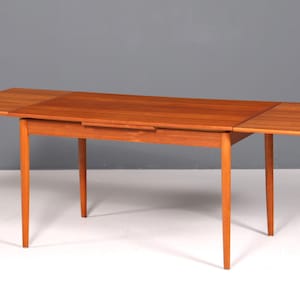 Puede incluir: Una mesa de comedor de madera de estilo moderno de mediados de siglo, con una tapa rectangular y cuatro patas delgadas. La mesa tiene dos hojas desplegables, lo que aumenta la superficie. La madera tiene un tono marrón rojizo cálido. La mesa mide unos 152 cm de largo.