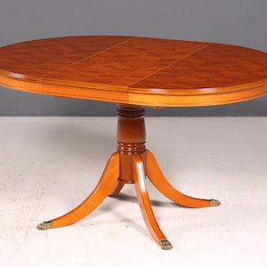Peut inclure: Une table à manger ovale en bois avec un plateau en placage brun riche présentant un motif géométrique. La table est soutenue par une colonne centrale qui se divise en quatre pieds incurvés, chacun terminé par des pieds en métal en forme de griffe.