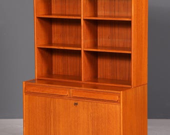 Peut inclure: Bibliothèque en bois de style milieu du siècle, de couleur brun rougeâtre chaleureux. La partie supérieure est composée d'étagères ouvertes, tandis que la partie inférieure comprend un meuble avec deux tiroirs et une porte. La bibliothèque repose sur quatre pieds fins.