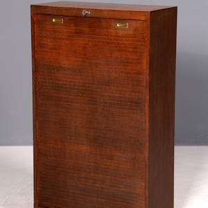 Könnte beinhalten: Ein hoher Holzschrank mit dunkelbraunem Finish. Der Schrank hat eine Rolltür mit horizontalen Lamellen und ein Schloss. Die Oberseite hat Messingbeschläge. Der Schrank ist ein Aufbewahrungsmöbel.