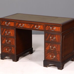 Op de afbeelding: Een bureau van donker hout met een rechthoekig blad en een groene lederen inleg. Het bureau heeft negen lades verdeeld over twee kolommen en een centrale lade, allemaal voorzien van messing handgrepen en een zichtbare houtnerfstructuur.