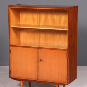 Könnte beinhalten: Ein Mid-Century-Modern-Holzschrank mit einem oberen Teil mit Glastüren und einem unteren Teil mit Massivtüren. Der Schrank hat zwei Regale hinter den Glastüren und steht auf vier konischen Beinen. Das Holz hat einen warmen, rotbraunen Farbton.