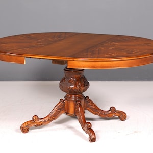 Puede incluir: Una mesa de comedor ovalada de madera con un acabado marrón intenso y patrones incrustados complejos en la superficie. La mesa cuenta con una base de pedestal central con detalles florales tallados y cuatro patas curvas con pies en voluta.