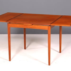 Könnte beinhalten: Ein Holztisch im Mid-Century-Modern-Stil mit rechteckiger Tischplatte und vier konischen Beinen. Der Tisch hat eine warme, rotbraune Oberfläche und verfügt über zwei Klappblätter, die die Oberfläche verlängern. Der Tisch ist ca. 76 cm lang.