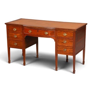 Op de afbeelding: Een mahoniehouten bureau met een rechthoekig blad voorzien van een lederen inleg. Het bureau heeft een centrale lade geflankeerd door twee kolommen van drie laden, elk met ovale koperen handgrepen. Het meubel staat op vier taps toelopende poten.