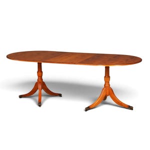 Pode incluir: Uma mesa de jantar oval de madeira com um acabamento castanho quente. O tampo da mesa apresenta uma costura visível no centro. É suportada por dois pedestais centrais, cada um ramificando-se em três pernas curvas com rodízios.