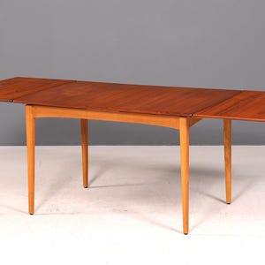 Peut inclure: Table de salle à manger de style moderne du milieu du siècle, avec un plateau rectangulaire extensible. Le plateau est de couleur marron chaud et les pieds sont d'une teinte bois naturel plus claire. La table est présentée avec les rallonges déployées.