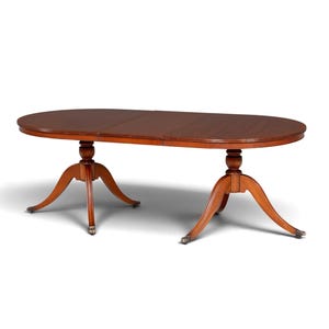 Op de afbeelding: Een ovale houten eettafel met een gladde, donkerbruine afwerking. De tafel heeft twee centrale zuilvoeten, elk met drie gebogen poten die eindigen in metalen klauwvoeten. Het tafelblad heeft een zichtbare naad die wijst op een uitschuifbaar ontwerp.