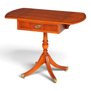 Pode incluir: Uma mesa de pedestal de madeira com acabamento castanho quente, apresentando um tampo retangular com duas abas laterais dobráveis, uma gaveta central com puxador oval de latão, um suporte de coluna torneado e uma base de tripé com rodízios.