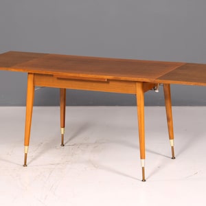 Peut inclure: Une table à manger rectangulaire en bois avec une finition brun chaud et un plateau extensible. La table présente quatre pieds fuselés, chacun accentué par une virole métallique dorée à la base, offrant une base stable à la surface plane.