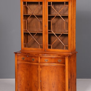 Könnte beinhalten: Ein hoher Holzschrank in hellbrauner Farbe mit zwei verglasten Türen, die im oberen Bereich ein geometrisches Gittermuster aufweisen. Der untere Teil besteht aus zwei Schubladen mit Metallgriffen über zwei massiven Holztüren mit gewölbter Front.