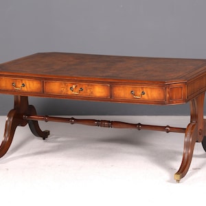 Puede incluir: Un escritorio rectangular de madera con una superficie marrón pulida y un borde oscuro. La parte frontal incluye tres cajones, cada uno con un tirador de latón. El escritorio se apoya sobre cuatro patas curvas unidas por un travesaño.