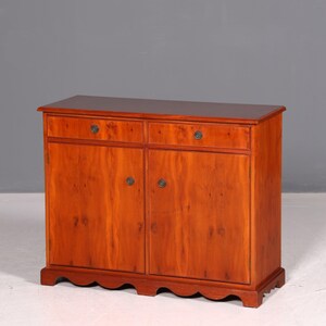 Könnte beinhalten: Ein hölzernes Sideboard mit einer warmen, orange-braunen Holzmaserung, zwei oberen Schubladen und zwei unteren Schranktüren. Jede Schublade und Tür ist mit einem kleinen, dunklen runden Knopf versehen. Der Sockel hat einen gewellten Rand.