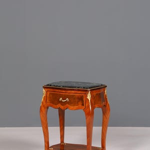 Op de afbeelding: Een kleine, antieke houten bijzettafel met een donker marmeren blad en een enkele lade. De tafel heeft sierlijke gouden accenten en gebogen poten. De lade heeft een kleine, gebogen handgreep. De tafel is ongeveer 61 cm hoog.