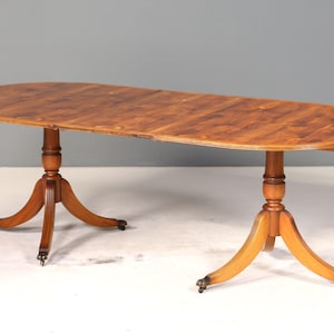 Può includere: Tavolo da pranzo ovale in legno con finitura marrone chiaro. Il tavolo ha due basi a piedistallo con tre gambe curve ciascuna e piccole rotelle. Il tavolo è lungo circa 183 cm.