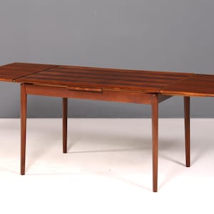 Pode incluir: Uma mesa de jantar retangular de madeira com um rico padrão de veios castanhos escuros. A mesa inclui duas abas extensíveis em cada lado, apoiadas por quatro pernas cónicas. A superfície apresenta um acabamento em folheado às riscas.