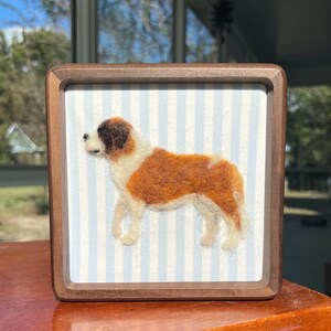 May include: A framed felted art piece featuring a Saint Bernard dog. The dog is depicted in shades of brown, white, and tan, set against a light blue and white striped background. The frame is made of dark wood.