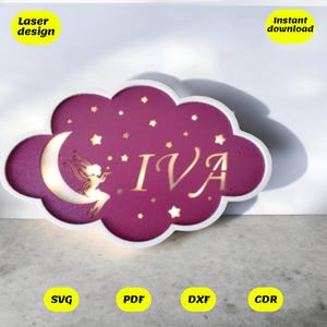 Peut inclure: Une veilleuse en bois en forme de nuage rose avec une découpe d'une fée assise sur un croissant de lune et le nom "IVA" au centre. La veilleuse est décorée de petites étoiles.