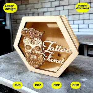 Può includere: Una scatola esagonale in legno con un design a laser di un teschio con barba e baffi, e le parole "Tattoo Fund". La scatola è su una superficie grigia. L'immagine include il testo "Laser design" e "Download immediato".