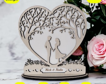 Può includere: Topper per torta nuziale in legno tagliato al laser a forma di cuore con un albero e due figure in piedi sotto. La base del topper ha il testo "Nick & Kaila" e una fila di cuori.