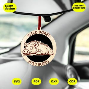 May include: A wooden car charm with a laser-cut design of a sleeping boar inside a black circle. The words "WILD BORED" are arched above and below the boar. It hangs from a red cord. The charm is suspended from a car's rearview mirror.