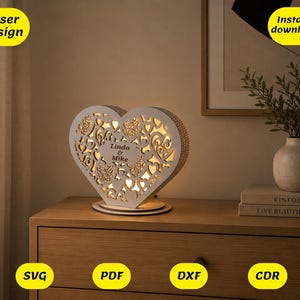 May include: A heart-shaped wooden lamp, laser-cut with floral and scrollwork patterns. The lamp has the names "Linda & Mike" and emits a warm light. It is displayed on a wooden surface.