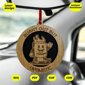 May include: A wooden ornament with a black circle in the center. The circle has a cartoon image of a burnt marshmallow with a smiling face and the text "Burnt Out But Optimistic".