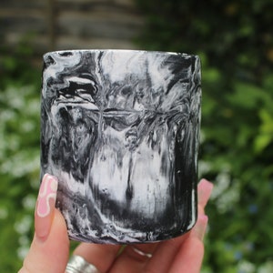 May include: A cylindrical container with a black and white marbled design. The swirling pattern creates an abstract effect. The container is held in a hand with a pink and white nail design, against a blurred green background.