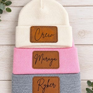 May include: Three knit beanies stacked vertically in cream, light pink, and gray. Each beanie features a rectangular brown leather patch centered on the cuff, engraved with the names Crew, Maraya, and Ryker in a black cursive script font.