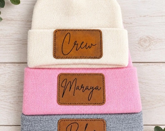 May include: Three knit beanies stacked vertically in cream, light pink, and gray. Each beanie features a rectangular brown leather patch centered on the cuff, engraved with the names Crew, Maraya, and Ryker in a black cursive script font.