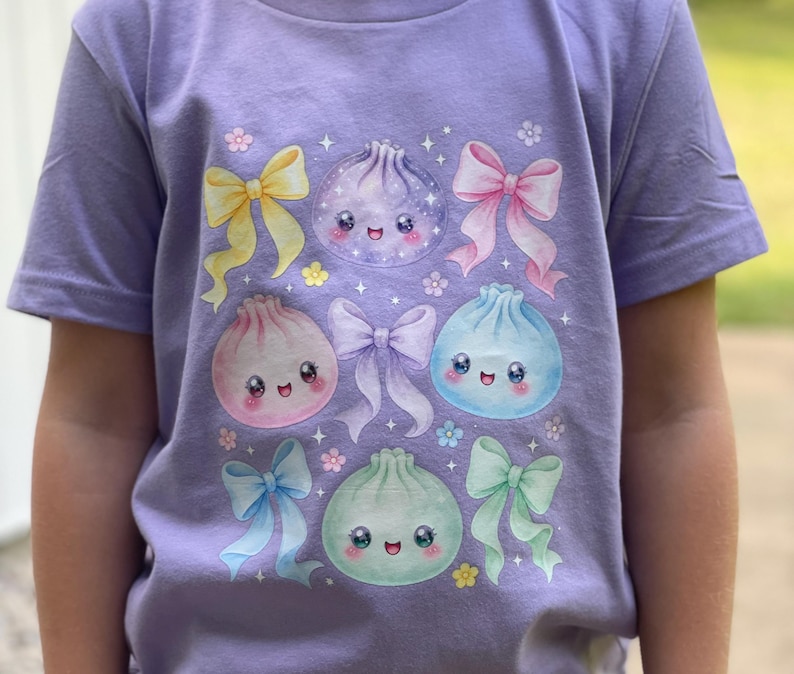 Może przedstawiać: Jasnofioletowy t-shirt z kr&oacute;tkim rękawem z nadrukiem graficznym przedstawiającym cztery okrągłe, uśmiechnięte postacie pierożk&oacute;w w kolorach fioletowym, r&oacute;żowym, niebieskim i zielonym, przeplatane pięcioma pastelowymi kokardkami oraz małymi białymi gwiazdkami.