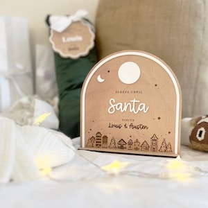 May include: A wooden sign with a crescent moon and a star, reading "Sleeps until Santa visits Louis & Austen". The sign is decorated with a small town silhouette and is sitting on a white bed with a white blanket and string lights.