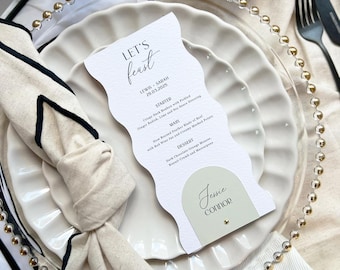 May include: A white card with black text on a white plate with a gold beaded rim. The card has a menu for a wedding reception with the text "LET'S feast" at the top. The menu includes a starter, main course, and dessert. The card also has a place card with the name "Jessie Connor" on it.
