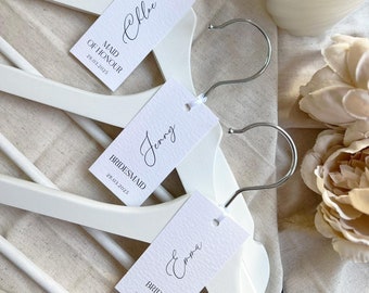 May include: Three white wooden hangers with white paper tags. The tags have the names Chloe, Jenny, and Emma written in cursive. The tags also have the titles Maid of Honour, Bridesmaid, and Bride, along with the date 29.03.2023.