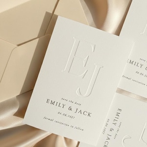 Pode incluir: Cartões de convite de casamento em marfim com iniciais em relevo e o texto "EMILY & JACK" e a data "06.06.2027". Os cartões estão dispostos sobre uma superfície de cetim, com um envelope visível no fundo.