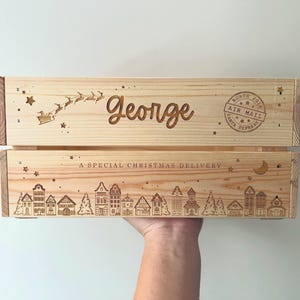 May include: A light-colored wooden crate featuring laser-engraved designs including a flying sleigh, the name George, a North Pole Air Mail stamp, a crescent moon, stars, and a row of houses with the text A SPECIAL CHRISTMAS DELIVERY.