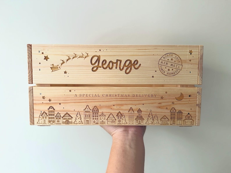 May include: A light-colored wooden crate featuring laser-engraved designs including a flying sleigh, the name George, a North Pole Air Mail stamp, a crescent moon, stars, and a row of houses with the text A SPECIAL CHRISTMAS DELIVERY.
