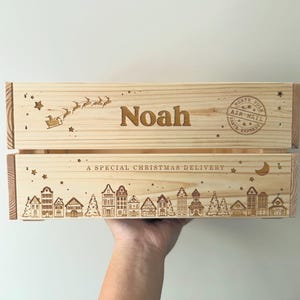 May include: A light-colored wooden crate featuring laser-engraved designs. The top panel displays the name Noah, a flying sleigh with reindeer, and a North Pole Air Mail stamp. The bottom panel shows a village scene with houses, trees, stars, a crescent moon, and the text A SPECIAL CHRISTMAS DELIVERY.