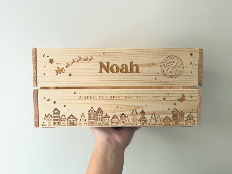 May include: A light-colored wooden crate featuring laser-engraved designs. The top panel displays the name Noah, a flying sleigh with reindeer, and a North Pole Air Mail stamp. The bottom panel shows a village scene with houses, trees, stars, a crescent moon, and the text A SPECIAL CHRISTMAS DELIVERY.