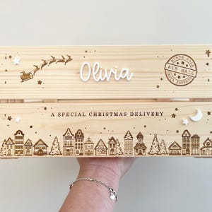 May include: A light-colored wooden crate featuring a laser-engraved North Pole air mail stamp, a silhouette of Santa in a sleigh, and a village scene. The text reads Olivia and A SPECIAL CHRISTMAS DELIVERY, accented with small white star and moon shapes.
