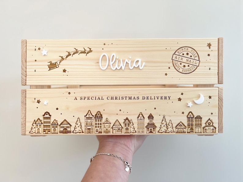 May include: A light-colored wooden crate featuring a laser-engraved North Pole air mail stamp, a silhouette of Santa in a sleigh, and a village scene. The text reads Olivia and A SPECIAL CHRISTMAS DELIVERY, accented with small white star and moon shapes.
