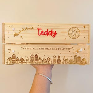 May include: A light-colored wooden crate featuring the red acrylic name Teddy, a laser-engraved silhouette of Santa in a sleigh, a North Pole Air Mail stamp, and the text A SPECIAL CHRISTMAS EVE DELIVERY above a detailed village scene.