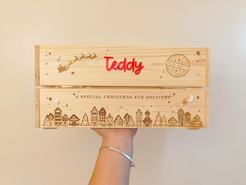 May include: A light-colored wooden crate featuring the red acrylic name Teddy, a laser-engraved silhouette of Santa in a sleigh, a North Pole Air Mail stamp, and the text A SPECIAL CHRISTMAS EVE DELIVERY above a detailed village scene.