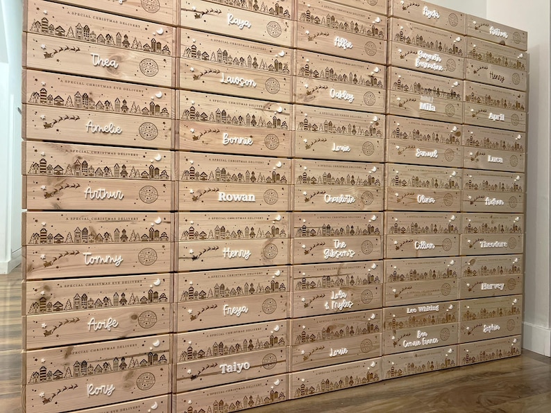 May include: A stack of rectangular light-colored wooden crates, each featuring a laser-engraved winter village scene with a flying sleigh and the text A SPECIAL CHRISTMAS DELIVERY. Each crate is personalized with a white script name attached.