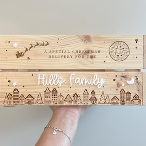 May include: A light-colored wooden crate featuring laser-engraved designs of a flying sleigh, a North Pole Air Mail stamp, and a village scene. The text reads A SPECIAL CHRISTMAS DELIVERY FOR THE Hills family, accented with small white stars and a moon.
