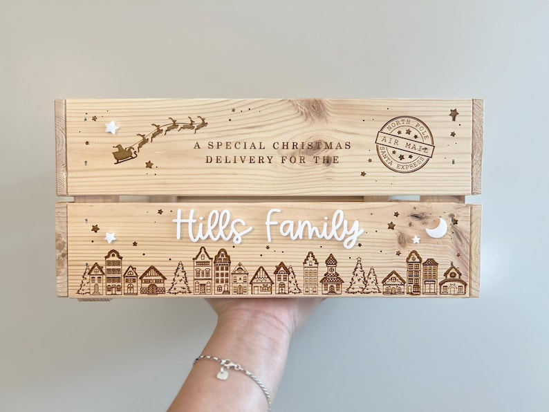 May include: A light-colored wooden crate featuring laser-engraved designs of a flying sleigh, a North Pole Air Mail stamp, and a village scene. The text reads A SPECIAL CHRISTMAS DELIVERY FOR THE Hills family, accented with small white stars and a moon.