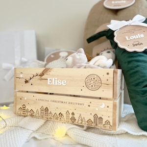 May include: A light-colored wooden crate featuring the name Elise, a silhouette of Santa in a sleigh, and a North Pole Air Mail stamp. The bottom panel displays a line drawing of a village with the text A SPECIAL CHRISTMAS DELIVERY.