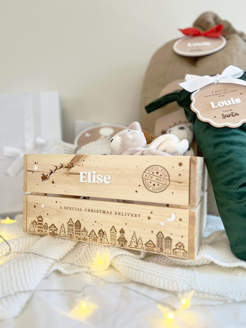 May include: A light-colored wooden crate featuring the name Elise, a silhouette of Santa in a sleigh, and a North Pole Air Mail stamp. The bottom panel displays a line drawing of a village with the text A SPECIAL CHRISTMAS DELIVERY.