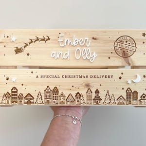 May include: A light-colored wooden crate featuring laser-engraved designs of a flying sleigh, a North Pole Air Mail stamp, and a village scene. The text reads Ember and Olly and A SPECIAL CHRISTMAS DELIVERY, accented with small white stars and a moon.