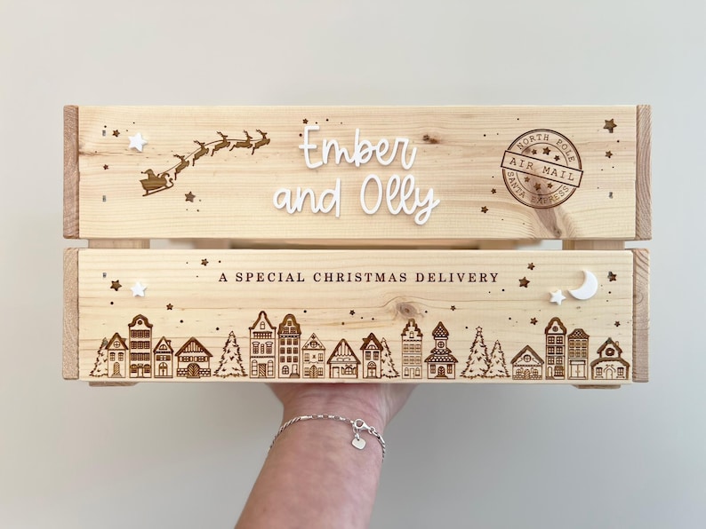 May include: A light-colored wooden crate featuring laser-engraved designs of a flying sleigh, a North Pole Air Mail stamp, and a village scene. The text reads Ember and Olly and A SPECIAL CHRISTMAS DELIVERY, accented with small white stars and a moon.